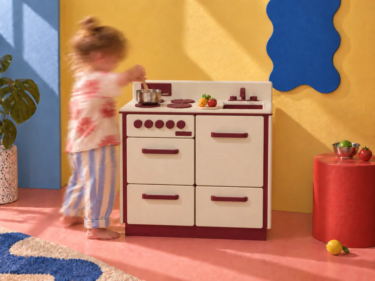 The bone and blood-red Little Range on a colourful yellow, blue and pink set, with a child blurred in motion while playing at the hob.