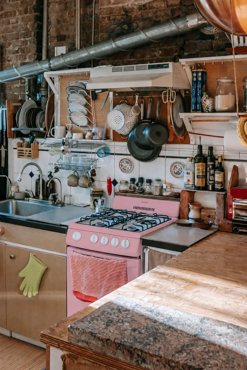 A kitchen full of hanging pots, warm timber and exposed brick, with the cooker recoloured dusty pink.