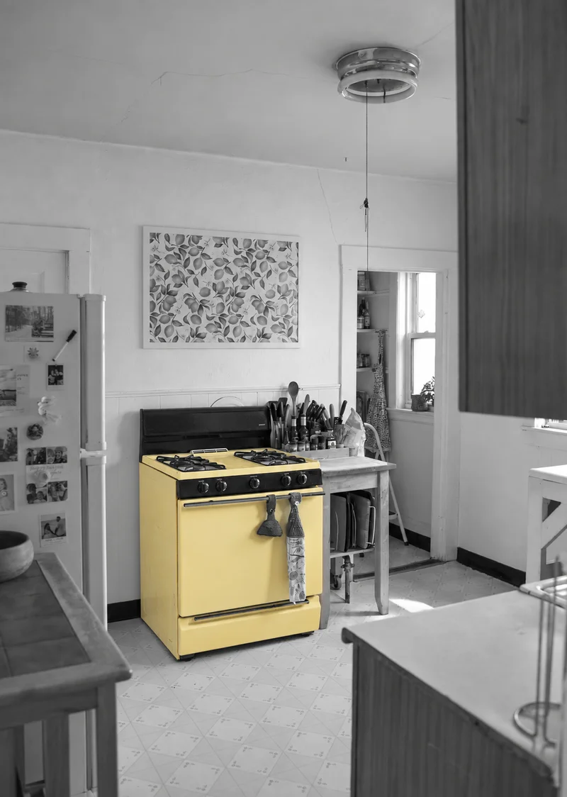 A butter-yellow cooker in a lived-in kitchen, with the surrounding room in black and white.