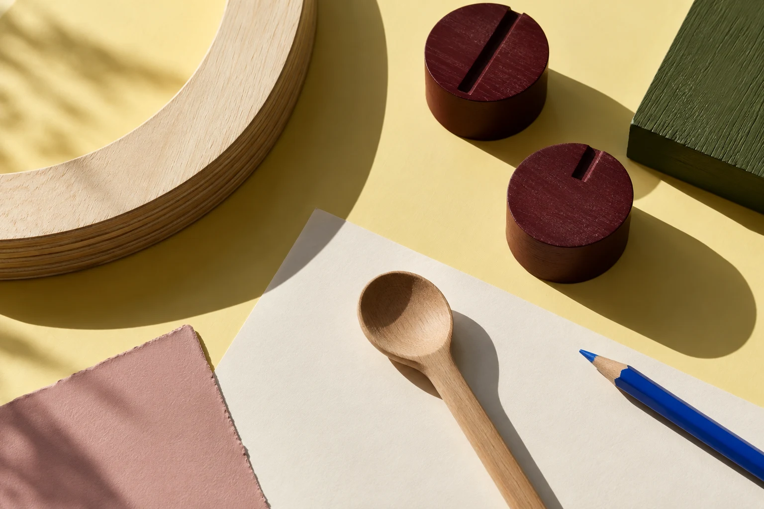 An editorial still life of plywood, colourful timber samples, wooden knobs and a spoon on a butter-yellow table.